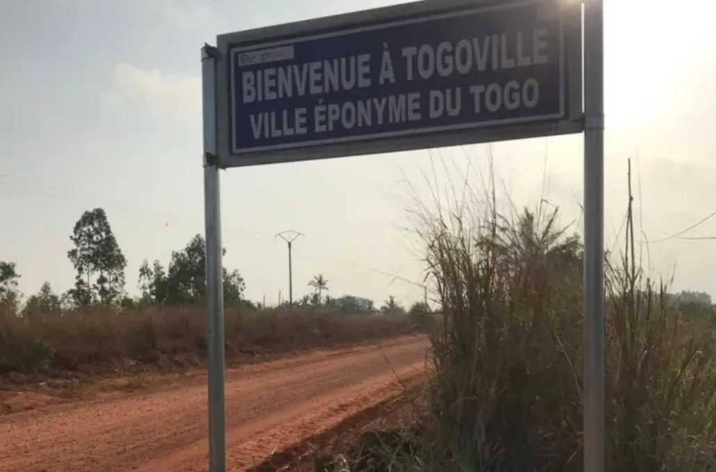 Une plaque indicatif sur laquelle est écrit : "Bienvenue à Togoville, ville éponyme du Togo". Elle est mise sur une piste en latérite avec la lumière du soleil au-dessus de la plaque. Située à environ 30 km de Lomé, la capitale togolaise, Togoville renferme pourtant des richesses culturelles qui peuvent être mises en valeur pour son développement | Photo: I.K. / BBC